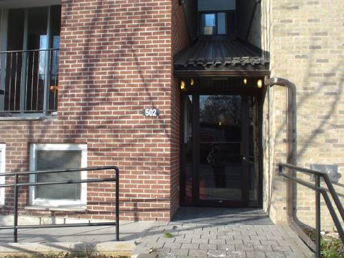 Building has recessed single door topped by angled corrugated eavestroughing, with iron banisters next to front brick steps and concrete wheelchair ramp