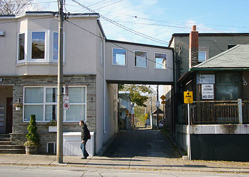 Grey two-storey house has catwalk with three windows that spans a driveway and connects it to a brick house