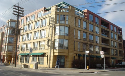 Five-storey building in buff brick occupies a corner lot, with green girders visible on a  roof terrace