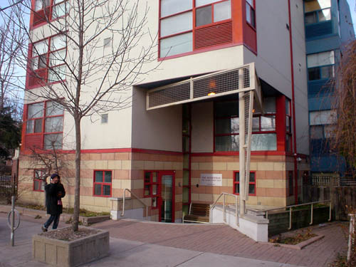 Cream-coloured building has red accents arounod windows, brown-and-tan banded brick at ground level, a sunken entrance with an angled metal grille at the entrance roof, and, at the rear, a light-blue additional wing