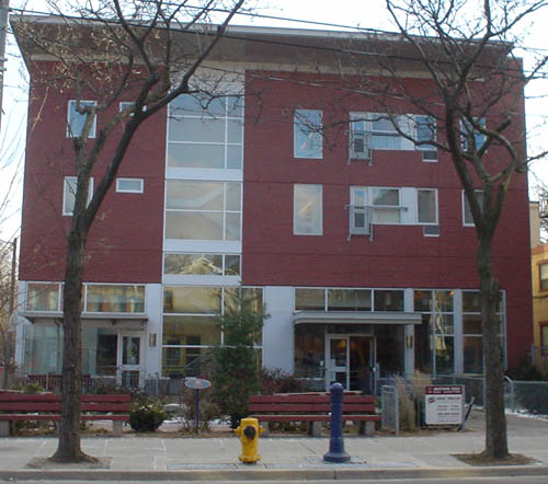 Three-storey building in red brick sits behind front courtyard with benches. Tiny street alongside has buff-brick building tucked away in back