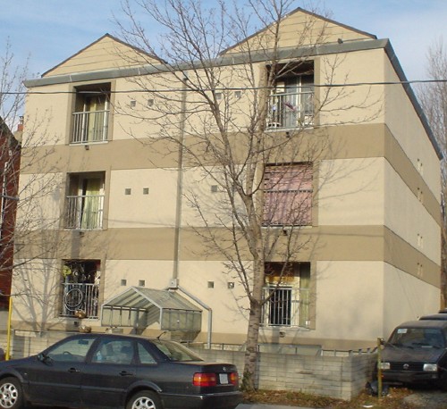 Buff-brick three-storey building has six front windows with grated railings and is divided into two halves with peaked roofs. Below the second and third storeys is a band of darker brick