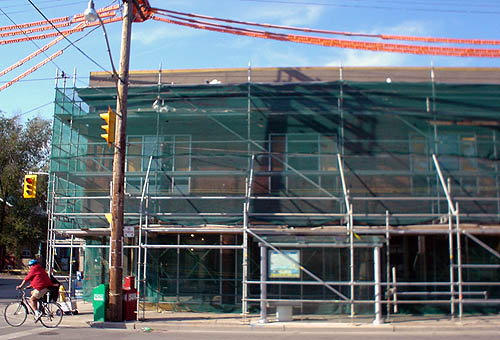 Glass windows are just barely visible behind green netting hanging down from construction scaffolding